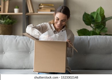 Smiling satisfied young woman customer sit on sofa unpack package open parcel, happy girl consumer holding cardboard box receive good online shop purchase at home, post mail shipping delivery concept