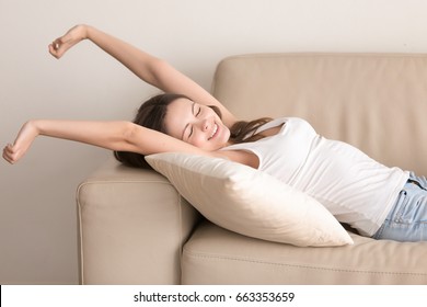Smiling satisfied young woman stretching while lying on comfortable sofa. Attractive lady waking up after pleasant day sleep, basking on couch with pillow. Female relaxing and feeling lazy on weekend