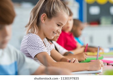 Smiling scholar girl sitting with other children in classroom and writing on textbook. Happy student doing homework at elementary school. Young schoolgirl feeling confident while writing on notebook. - Powered by Shutterstock