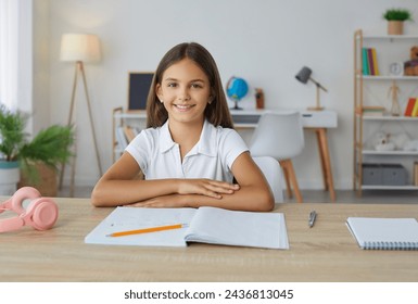 Smiling school girl sitting at desk doing her homework at home. Portrait of happy preteen student girl sitting at table with folded hands and smiling at camera. School education, homeschooling concept - Powered by Shutterstock