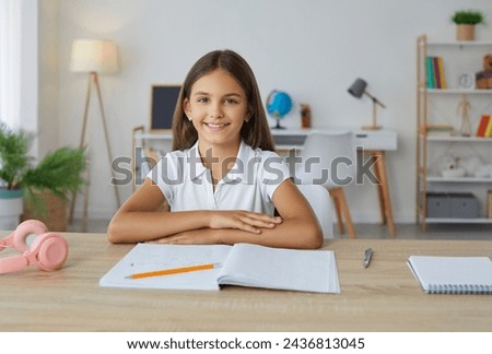 Smiling school girl sitting at desk doing her homework at home. Portrait of happy preteen student girl sitting at table with folded hands and smiling at camera. School education, homeschooling concept