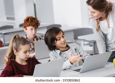 smiling schoolboy pointing at laptop near teacher in classroom