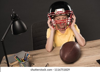 Smiling Schoolboy Putting On American Football Helmet At Table With Ball, Lamp, Colour Pencils And Books On Grey Background 