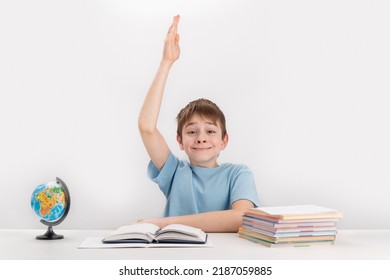 Smiling Schoolboy Raised His Hand During The Lesson. Excellent Student Knows The Answer. Portrait Of Student At Table Next To Notebooks.