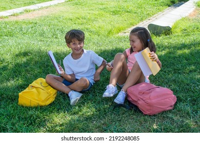 Smiling Schoolchildren Sitting On The Grass In The Schoolyard. Girl And Boy Doing Homework At School Recess. Back To School. Elementary School Children	