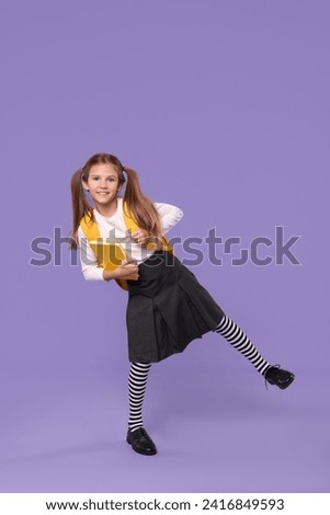 Smiling schoolgirl with backpack and book on violet background