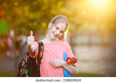 Smiling schoolgirl with backpack and skateboard showing thumbs up in sunlight. Educational course for teens. Back to school concept. Selective soft focus. - Powered by Shutterstock