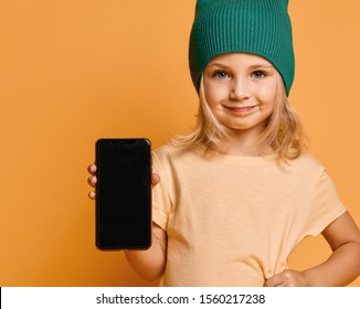 Smiling Screaming Little Girl Kid In Green Modern Winter Hat Showing Blank Screen Of New Popular Mobile Phone On Light Yellow Background