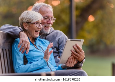 Smiling Senior Active Couple Sitting On The Bench Looking At Tablet Computer. Using Modern Technology By Elderly.