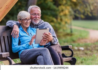 Smiling Senior Active Couple Sitting On The Bench Looking At Tablet Computer. Using Modern Technology By Elderly.