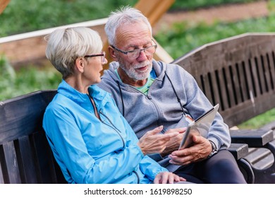 Smiling Senior Active Couple Sitting On The Bench Looking At Tablet Computer. Using Modern Technology By Elderly.