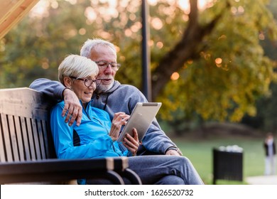 Smiling senior active couple sitting on the bench looking at tablet computer. Using modern technology by elderly.