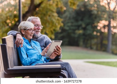 Smiling Senior Active Couple Sitting On The Bench Looking At Tablet Computer. Using Modern Technology By Elderly.