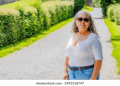 Smiling senior adult hiker standing on a rural road, looking to camera, green bushes and path disappearing in blurred background, casual clothes, jeans, white blouse and sunglasses, sunny summer day - Powered by Shutterstock