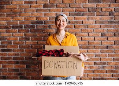 A Smiling Senior Asian Female Volunteer Holds A Cardboard Box With A Donation, For Reuse Or Recycle Of Old Clothes. Portrait Of A Happy Woman Stands Against Brick Wall With Donation Box