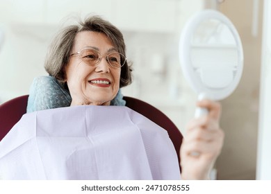 Smiling senior attractive female patient holding mirror looking at teeth while sitting in dental chair in modern dentistry. Concept of dental care, health care - Powered by Shutterstock