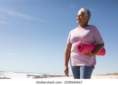 Smiling senior biracial woman holding pink yoga mat while looking away at beach against blue sky. active lifestyle and fitness. - Powered by Shutterstock