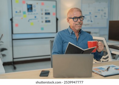 Smiling senior businessman enjoys a coffee break at his desk, reading notes in a relaxed and productive work environment - Powered by Shutterstock