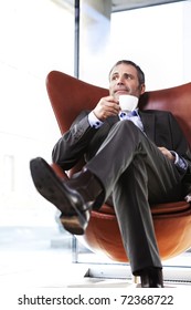 Smiling senior businessman in grey suit sitting in red office chair looking outside and enjoying a cup of coffee with bright background.