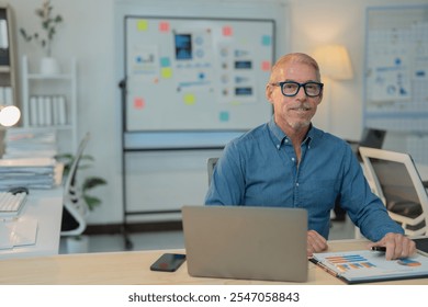 Smiling senior businessman sitting at his desk working on laptop and analyzing financial charts and graphs in the office - Powered by Shutterstock