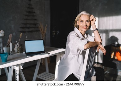 Smiling Senior Caucasian Female Entrepreneur Sitting On Chair And Looking At Camera. Concept Of Small Business And Entrepreneurship. Modern Successful Woman At Dark Art Studio With Sunny Lighting