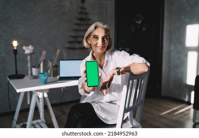 Smiling Senior Caucasian Female Entrepreneur Sitting On Chair And Showing Smartphone With Green Screen. Small Business And Entrepreneurship. Modern Successful Woman At Dark Art Studio With Lighting