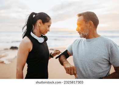 Smiling senior caucasian lady in sportswear and headphones use phone app on man phone, enjoy training on beach in morning, outdoor. Relationship, device for cardio sports together - Powered by Shutterstock