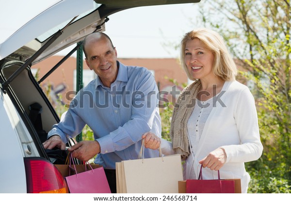 Smiling senior couple with bags near car at shopping\
center parking lot