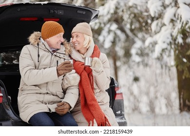 Smiling senior couple enjoying cup of hot coco outdoors in winter while sitting in car trunk, copy space - Powered by Shutterstock