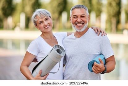 Smiling senior couple with exercise mats standing at park, positive mature man embracing his elderly wife after fitness or yoga class in nature. Wellness and healthy lifestyle on retirement