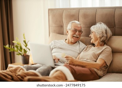 Smiling senior couple with laptop shopping online on the internet and looking at each other while lying down in bed. - Powered by Shutterstock