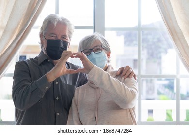 A Smiling Senior Couple Look At Camera Wearing Surgical Protective Mask, Making Hear Shape With Their Hands - In Lockdown At Home Due To The Coronavirus Infection