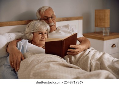 Smiling senior couple reading book together while relaxing in bedroom. - Powered by Shutterstock