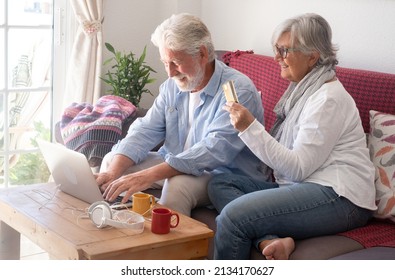 Smiling Senior Couple Shopping Online By Laptop Sitting On Sofa At Home. Old Attractive Gray Haired People Pay With Credit Card, Bank, Consumerism Concept