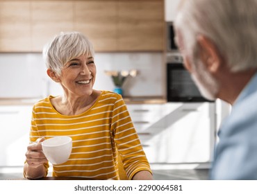 Smiling senior couple sitting at dining table in front of kitchen cabinets, drinking coffee and talking at home - Powered by Shutterstock