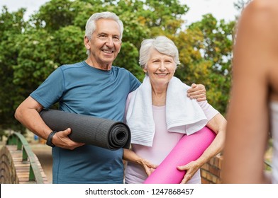 Smiling Senior Couple Standing And Holding Yoga Mat At Park. Old Man Embrace Her Elderly Wife As They Are Ready For Yoga Practice. Retired Man And Woman Doing Fitness Outdoor With Coach.