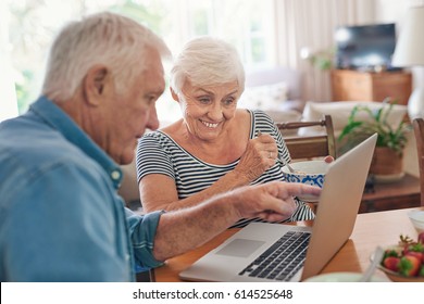 Smiling Senior Couple Talking Together Over A Laptop While Sitting At Their Dining Table In The Morning Eating A Healthy Breakfast