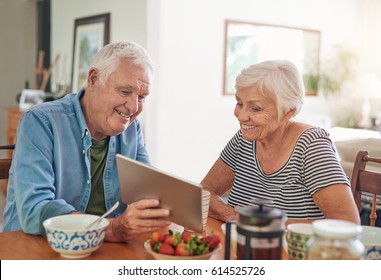 Smiling Senior Couple Talking Together Over A Digital Tablet While Sitting At Their Dining Table In The Morning Eating A Healthy Breakfast