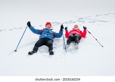 Smiling Senior Couple Walking With Nordic Walking Poles In Snowy Winter Forest. Elderly Wife And Husband Lying On Snow Outdoors And Happy Laugh. Active Lifestyle After Retirement Concept.