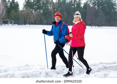 Smiling Senior Couple Walking With Nordic Walking Poles In Snowy Winter Park. Elderly Wife And Husband Doing Healthy Exercise Outdoors. Active Lifestyle Concept.