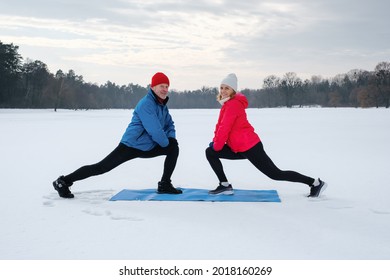 Smiling Senior Couple Warming Before Warms Up And Does Stretching Before Workout On Snowy Winter Lake. Elderly Wife And Husband Doing Fitness Outdoors. Active Lifestyle Concept.