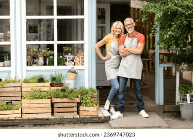 Smiling Senior Couple Wearing Aprons Posing Outdoors Near Their Greenhouse Bar, Happy Mature Couple Small Business Owners Standing Outside At Terrace And Looking At Camera, Free Space - Powered by Shutterstock