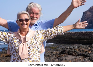 Smiling senior couple with white hairs having fun at the beach with raised arms - horizon over water - concept of active lifestyle for two elderly people in holidays - Powered by Shutterstock