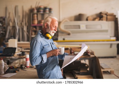 Smiling senior craftsman is casually reading blueprints and holding a cup of tea in his workshop - Powered by Shutterstock