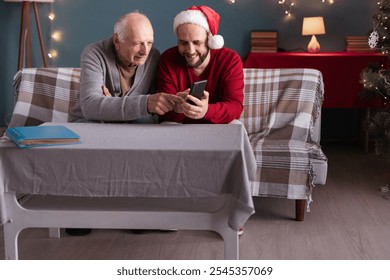 Smiling senior father and adult son in Santa hat explore smartphone together, creating joyful memories on Christmas. Technology and family bonding in cozy holiday atmosphere at home. - Powered by Shutterstock