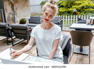 Smiling senior female blogger in casual wear sitting in coffee shop using laptop and smartphone.