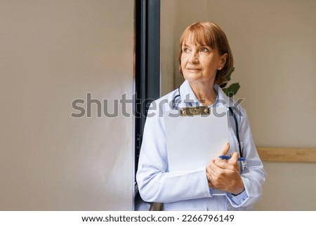 Smiling senior female doctor with lab coat in her office holding a clipboard with medical records, she is not looking at camera.