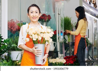 Smiling Senior Flower Shop Owner Standing With Bucket Of Fresh White Roses, Her Coworker Checking Flowers In Glass Indow In Background