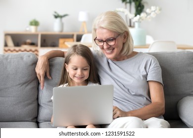 Smiling Senior Grandma And Cute Little Kid Granddaughter Watching Cartoons On Laptop Together, Happy Older Grandmother Playing Game Or Shopping Ordering Buying Online With Child Girl On Computer