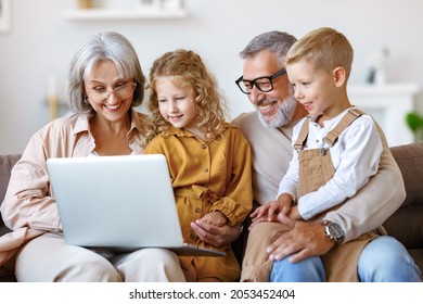 Smiling Senior Grandparents And Cute Little Grandchildren Boy And Girl Looking At Laptop Computer Screen While Sitting On Sofa At Home, Happy Kids Watching Cartoons With Grandpa And Grandma On Weekend